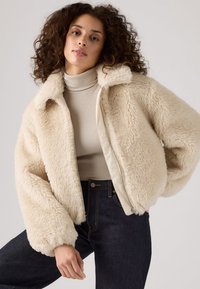 Cremefarvet jakke i imiteret pels med høj krave, lynlås og oversize ærmer. Båret over en lys rullekrave og mørke jeans.