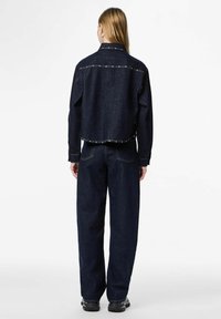 Mørk denimjakke med synlige sømmer og metalliske detaljer, kombinert med avslappede mørke jeans og svarte sko. Sett bakfra.