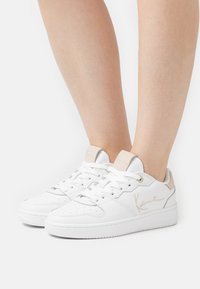 Des baskets blanches en cuir présentent une texture lisse, un bout arrondi, des accents beiges sur le talon, et un logo discret sur le côté et la languette.