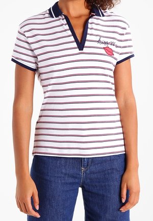 Kobieta w białej koszulce polo z cienkimi czerwonymi i granatowymi poziomymi paskami, granatowym kołnierzykiem oraz haftem ust i kursywnego tekstu na piersi.