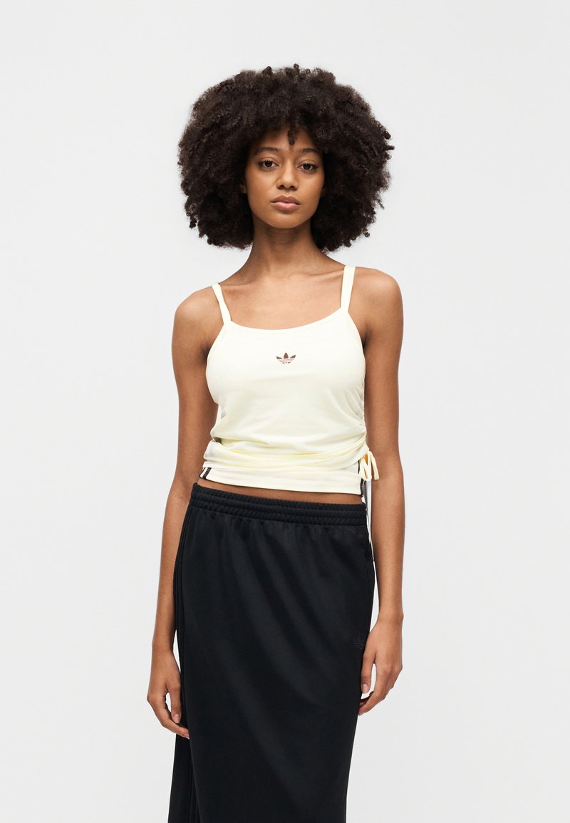 Haut jaune sans manches avec liens ajustables sur les côtés, arborant un petit logo marron au centre, associé à une jupe noire.