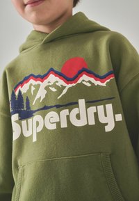 Zelená mikina s kapucí s předním potiskem zobrazujícím hory, stromy a červené slunce. Pod designem je vytištěno "Superdry" tučným bílým textem.