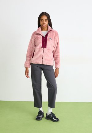 Vaaleanpunainen fleece-takki, jossa on maroon-värinen vetskari edessä, sivutaskut ja korkea kaulus, yhdistettynä harmaisiin housuihin ja mustiin lenkkareihin.