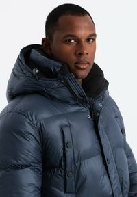 Giubbotto imbottito blu navy con cappuccio rimovibile, dotato di tasche con zip e bottone a pressione, realizzato in tessuto lucido e morbido con un rivestimento interno soffice.