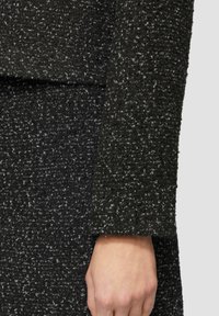 Detail eines Kleidungsstücks aus schwarzen und grauen strukturierten Stoffen, mit grober Webart, gekürztem Ärmel und sichtbaren Nähten entlang der Kante.