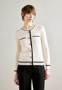 Cardigan en tricot blanc avec des bords noirs, présentant un col rond, deux poches à l'avant et des boutons noirs au centre. Porté sur un pantalon noir.