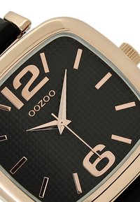 Montre carrée noire avec des accents dorés, cadran texturé, grands chiffres 12 et 6, fins aiguilles des heures et des minutes, et un bracelet en cuir noir.