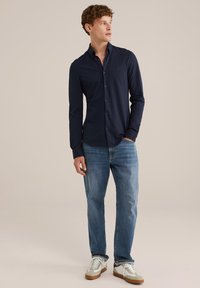 Tamsiai mėlyna ilgomis rankovėmis su sagutėmis marškiniai laisvo silueto, derinami su šviesiai mėlynais džinsais ir baltais sportbačiais su pilkomis ir smėlio spalvos detalėmis.