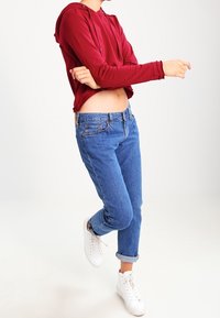 Sudadera corta roja con capucha, jeans azules de cintura alta y zapatillas blancas. El conjunto presenta texturas suaves y casuales, con un ajuste relajado.