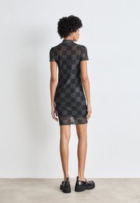Zwarte mesh jurk met korte mouwen en een geruit patroon met ronde logo's. Strakke pasvorm die tot halverwege de dij komt, gecombineerd met zwarte chunky schoenen.