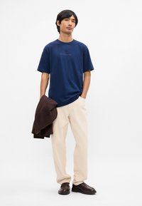 Μπλε βαθύ βαμβακερό μπλουζάκι με κοντά μανίκια, με μικρό λογότυπο. Συνδυάζεται με ανοιχτό μπεζ παντελόνι και κρατάει ένα καφέ μπουφάν. Σκοτεινά υποδήματα συμπληρώνουν την εμφάνιση.