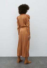 Abito lungo a maniche lunghe color marrone con pois bianchi, gonna plissettata e vita aderente. Indossato con stivaletti marroni alla caviglia, che mostrano una texture liscia e un design sartoriale.