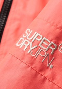 Koralfarvet jakke med gitterstruktur, med hvid broderet tekst "SUPER DRY JPN." Detaljeret syning synlig nær sømmen.