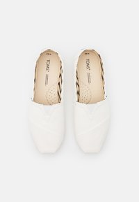 Hvit kanvas slip-on sko med V-formet design, glatt tekstur og svart mønstrete fôr. TOMS-logo synlig på sålen.
