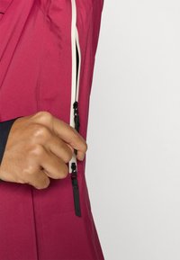 Fuchsiajakke med høj krave og kontrasterende hvid lynlås. Nærbillede af en hånd, der griber fat i lynlåstrækkeren, med en glat og jævn tekstur.