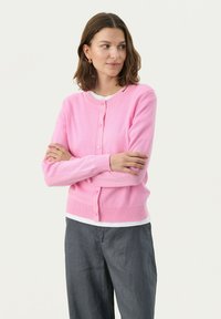 Gilet tricoté rose clair à manches longues, boutons sur le devant et ourlet côtelé ; porté par-dessus une chemise blanche avec un pantalon gris ample.