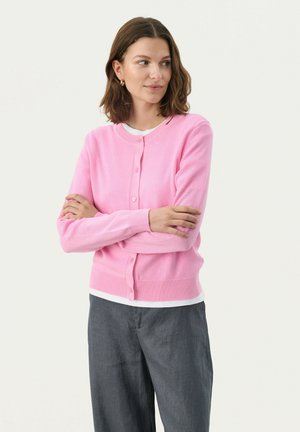 Hellrosa gestrickte Strickjacke mit langen Ärmeln, vorderen Knöpfen und geripptem Saum; getragen über einem weißen Hemd mit grauen, locker sitzenden Hosen.
