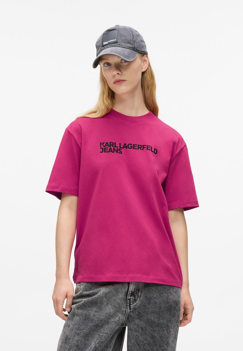 Fukszia színű pamut póló rövid ujjal, fekete "KARL LAGERFELD JEANS" felirattal. Szürke sapkával párosítva, amelyen egy téglalap alakú folt található.