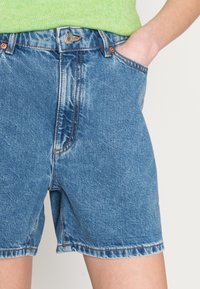 Persona che indossa pantaloni corti di jeans blu e una maglietta verde, con una mano comodamente appoggiata nella tasca anteriore dei pantaloni.