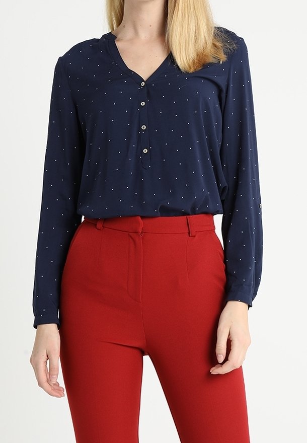 Femme portant une blouse bleu marine à manches longues avec des pois blancs et un pantalon rouge taille haute, debout sur un fond uni.