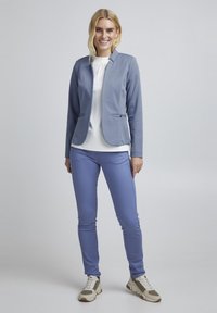 Světle modrý blazer s dvěma předními kapsami, nošený přes bílý top. Ladící světle modré kalhoty a tenisky s šedými akcenty. Hladká textura.