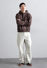 Sudadera marrón con un diseño a rayas, de corte holgado. Pantalones de pierna ancha en un tono beige claro, combinados con zapatillas oscuras con acentos blancos. Fondo neutro.