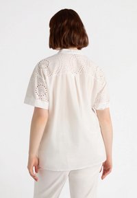 Camicia bianca a maniche corte con motivi a occhio di pernice sulle spalle, colletto classico e vestibilità rilassata, realizzata in un tessuto liscio e morbido.