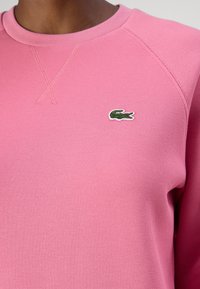 Sudadera texturizada rosa con cuello redondo y mangas raglán. Presenta un pequeño logo bordado de cocodrilo en verde y rojo en el pecho.