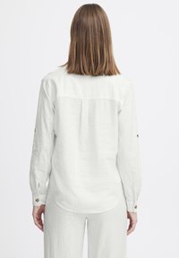 Chemise en lin blanc avec des manches longues. Présente un pli au dos et des accents de boutons sur les manches. Texture simple et lisse.