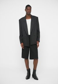 Dubbelknäppt kavaj med kritstrecksrand i mörkgrått, kombinerad med svarta shorts. Modellen bär ett vitt linne och svarta ankelboots.