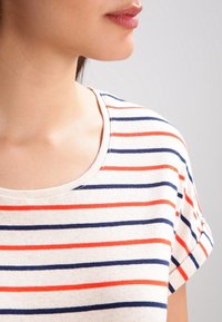 T-shirt à manches courtes rayé avec des rayures horizontales bleu marine et orange sur un fond crème, avec un col rond et une texture de tissu douce.
