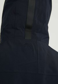 Granatowa, wodoodporna kurtka z fakturowanym kapturem, pionowym czarnym paskiem, haftowanym logo i wyraźnie zaznaczonymi szwami.