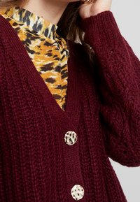 Burgundy knitted cardigan with a V-neck, featuring two large patterned buttons. Worn over a yellow, black, and white patterned top.