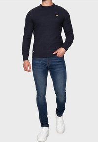 Marineblauwe gebreide trui met een ronde hals en een klein geel logo op de borst, gecombineerd met slim-fit jeans en witte schoenen.