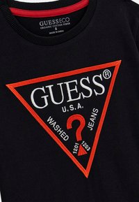 Černé tričko z organické bavlny s velkým trojúhelníkovým vyšitým logem v červené a bílé barvě. Text obsahuje "GUESS," "U.S.A," a "WASHED JEANS."