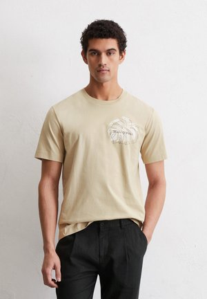 Beige cotton t-shirt with short sleeves, featuring a white abstract leaf design on the left chest. Paired with black trousers.