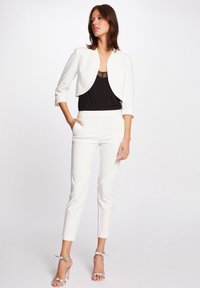 Weißer, kurzer Blazer über einem schwarzen Oberteil mit Spitze, kombiniert mit weißen, eng anliegenden Hosen und silbernen, hohen Sandalen.