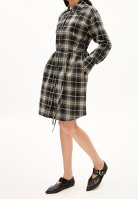 Abito camicia a quadri bianco e nero con vita regolabile, chiusura frontale con bottoni, maniche lunghe e tasche laterali. Indossato con ballerine nere.