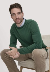 Grüner gestrickter Pullover mit rundem Ausschnitt, getragen über einem hellblauen Hemd mit Kragen. Slim-Fit-Cord-hosen in einem hellen Beigeton.