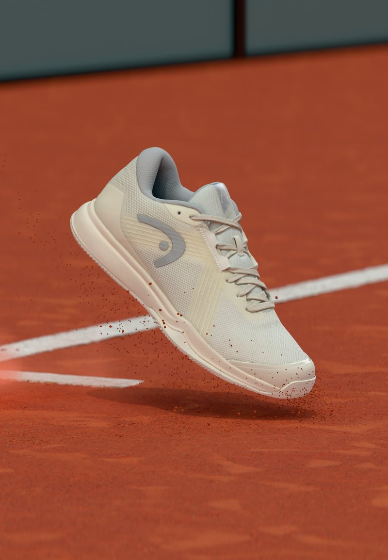 Zapato deportivo gris claro con parte superior texturizada, sistema de cordones y detalle de logotipo, flotando sobre una superficie de arcilla roja con partículas de polvo.