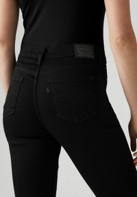 Jeans de mezclilla negra de talle alto, con bolsillos traseros y una etiqueta de marca elevada. Los acentos cosidos resaltan el diseño elegante.