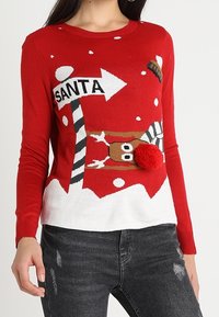 Pull en tricot rouge avec un design festif portant le texte "SANTA", un renne, des flocons de neige et un détail en pompon. Ourlet blanc.