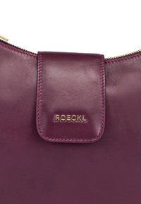 Structured leather pouch in deep purple with a curved top, featuring a gold zip, contrasting pink stitching, and a front flap with logo.