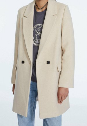 Beżowy wełniany płaszcz dwurzędowy noszony na ciemnej koszulce z grafiką i jasnoniebieskich dżinsach, z czarnym naszyjnikiem typu chokera.