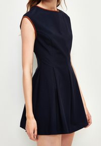 Marinblå ärmlös klänning med en figursydd överdel, vid kjol och kontrasterande brun kantning vid halsringningen och ärmhålen. Tillverkad av en bomullsblandning.