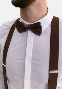 Pajarita de terciopelo marrón combinada con tirantes marrones, llevada sobre una camisa blanca de botones. La vista en primer plano resalta las texturas y colores.
