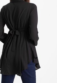 Blouse noire en tissu léger, dotée d'une ceinture à la taille, à manches longues et avec un ourlet fluide.