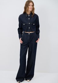 Tummansininen farkkutakki, jossa on hopeanväriset napit ja kaksi rintataskua, yhdistettynä leveälahkeisiin tummansinisiin farkkuihin. Pintakuvioinen kangas, rento tyyli.