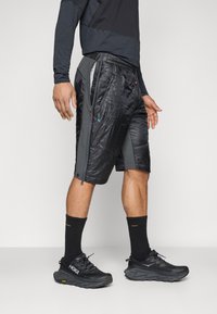Shorts noirs avec une texture brillante, panneaux latéraux gris et poches zippées. Portés avec des chaussures de sport noires et des chaussettes de sport assorties.
