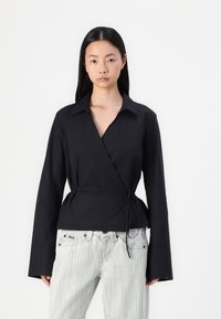 Black wrap shirt with long sleeves, a collar, and a tie at the waist. Paired with light striped jeans, creating a contrasting look.